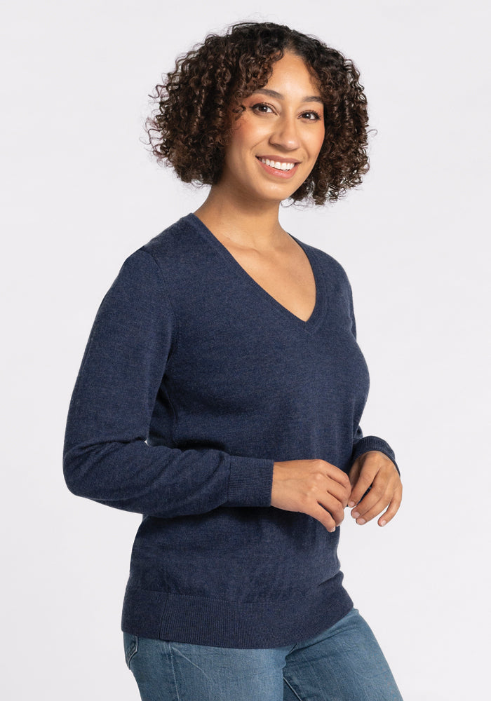 A woman with curly hair, wearing the Woolx Francesca Sweater in Twilight Storm and blue jeans, stands against a plain white background, smiling at the camera.