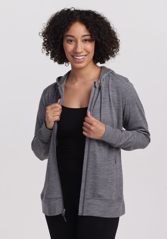 A person with curly hair wears the Woolx Ryann Hoodie - Graphite Heather over a black top, smiling against a white background. Known for its moisture-wicking properties, the lightweight travel hoodie is gripped by both hands. 