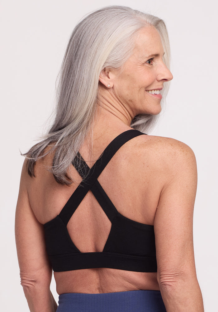 A smiling woman with long gray hair looks to the side, wearing a black Woolx Livi Bra with crisscross straps and blue leggings made from an odor-resistant Merino wool blend, set against a plain white background.