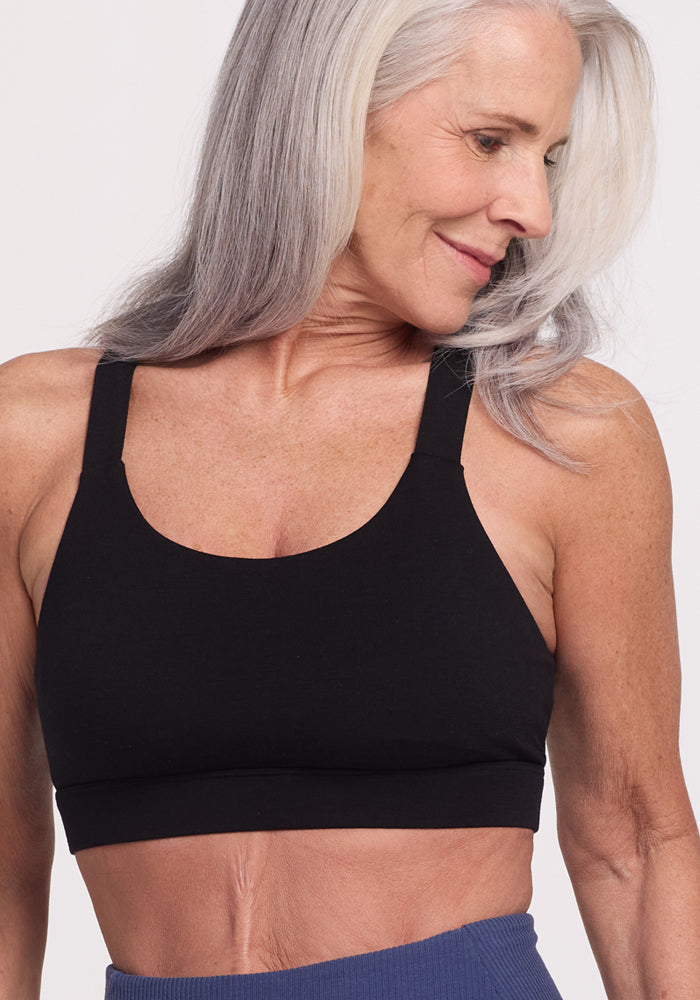 An older woman with long gray hair smiles while wearing the Woolx Livi Bra in black and blue leggings, posed against a white background. The sports bra features temperature-regulating fabric to ensure comfort. 