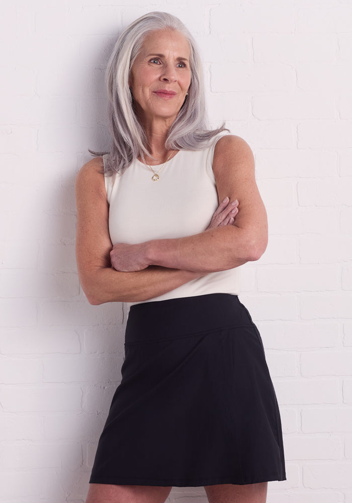 A woman with long gray hair stands against a white brick wall, smiling slightly with arms crossed and gazing to her right. She is wearing a sleeveless white top and the Topanga Skort in black by Woolx.