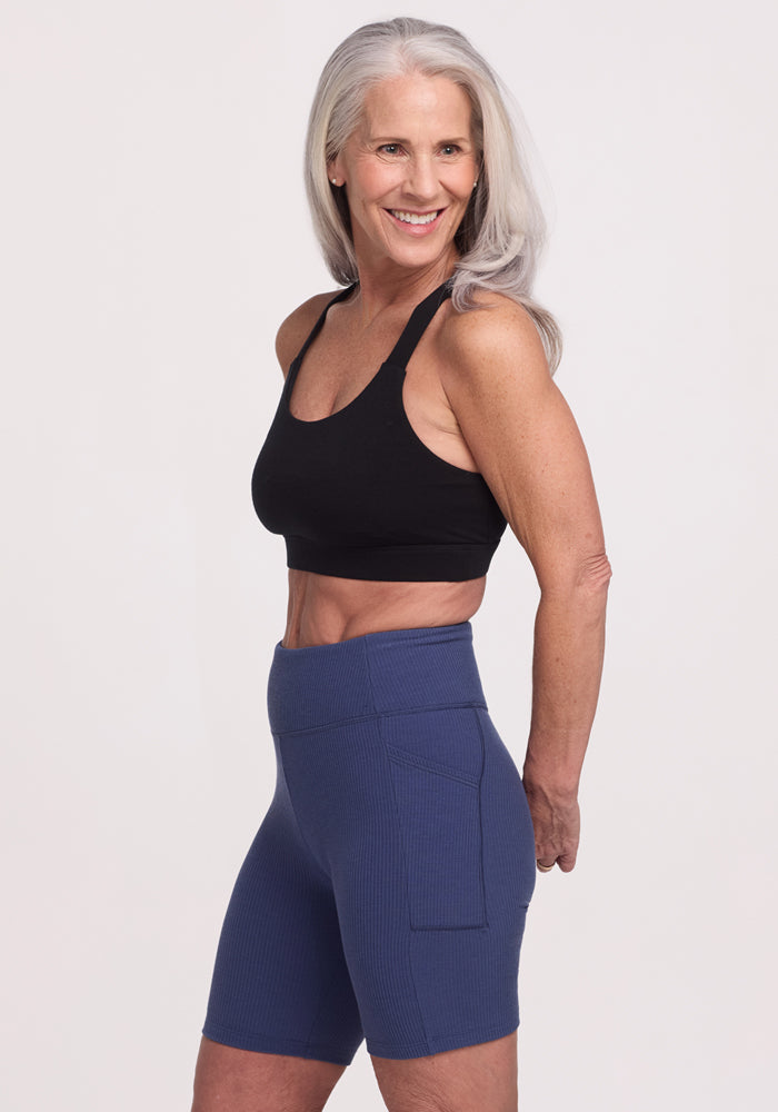 An older woman with long gray hair smiles while wearing the Woolx Livi Bra - Black and blue high-waisted shorts, crafted from an odor-resistant Merino wool blend, against a plain white background.