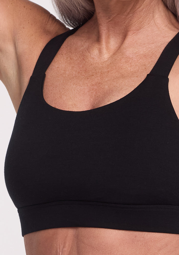 A close-up showcases a person wearing the Livi Bra - Black by Woolx, made from an odor-resistant Merino wool blend. They lift one arm slightly, accentuating a fit upper body against a plain background, emphasizing the bra and chest area.
