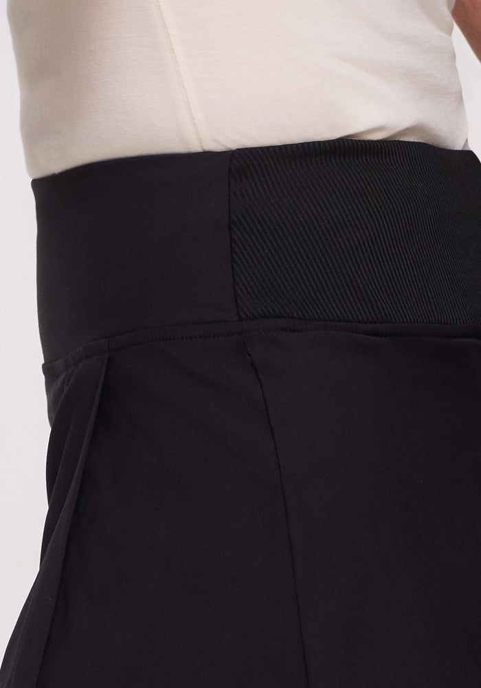 Close-up of a person wearing the Woolx Topanga Skort in black. A wide, textured waistband pairs with a white shirt visible above it. The skort features a side pleated detail.