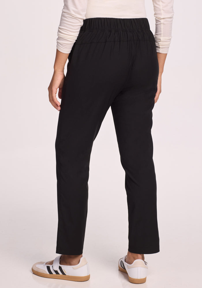 Someone wearing the Woolx Nicole Pants in black faces away from the camera, paired with a light long-sleeve top and white sneakers featuring black stripes, against a plain background.