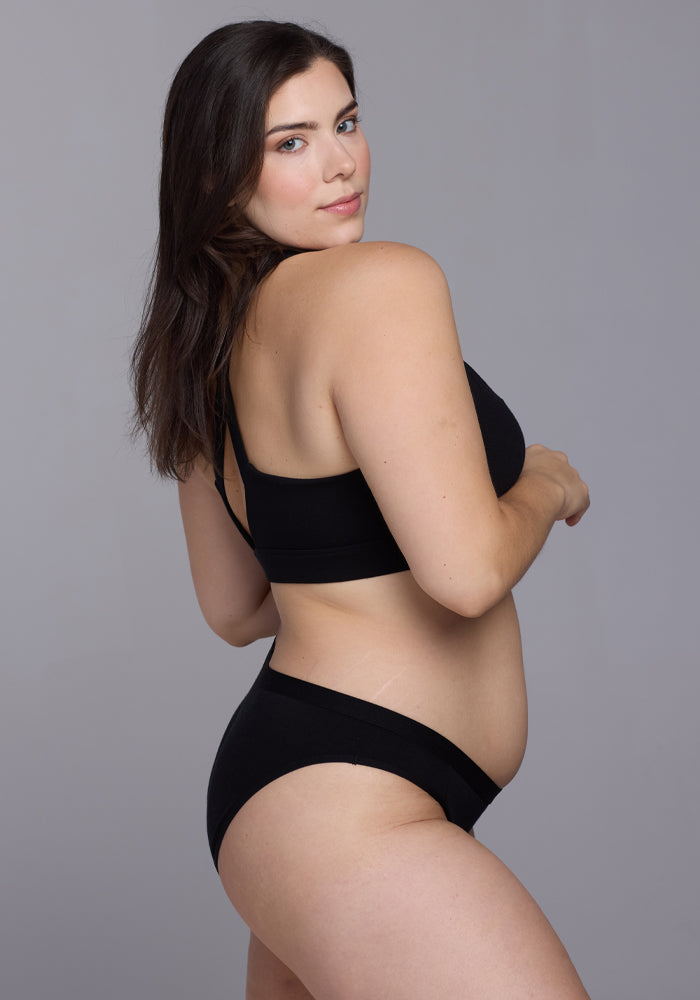 A woman with long dark hair confidently poses in Woolx's Roxie Bikini Underwear - Black, offering full back coverage, against a plain gray background while glancing over her shoulder.