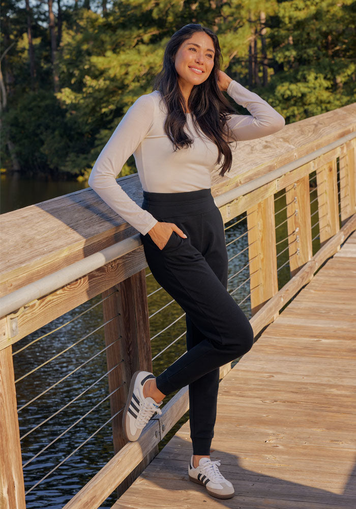 A woman in a white long-sleeve shirt, black Woolx Willa Joggers, and white sneakers smiles while leaning against a wooden bridge railing, with trees and water in the background.