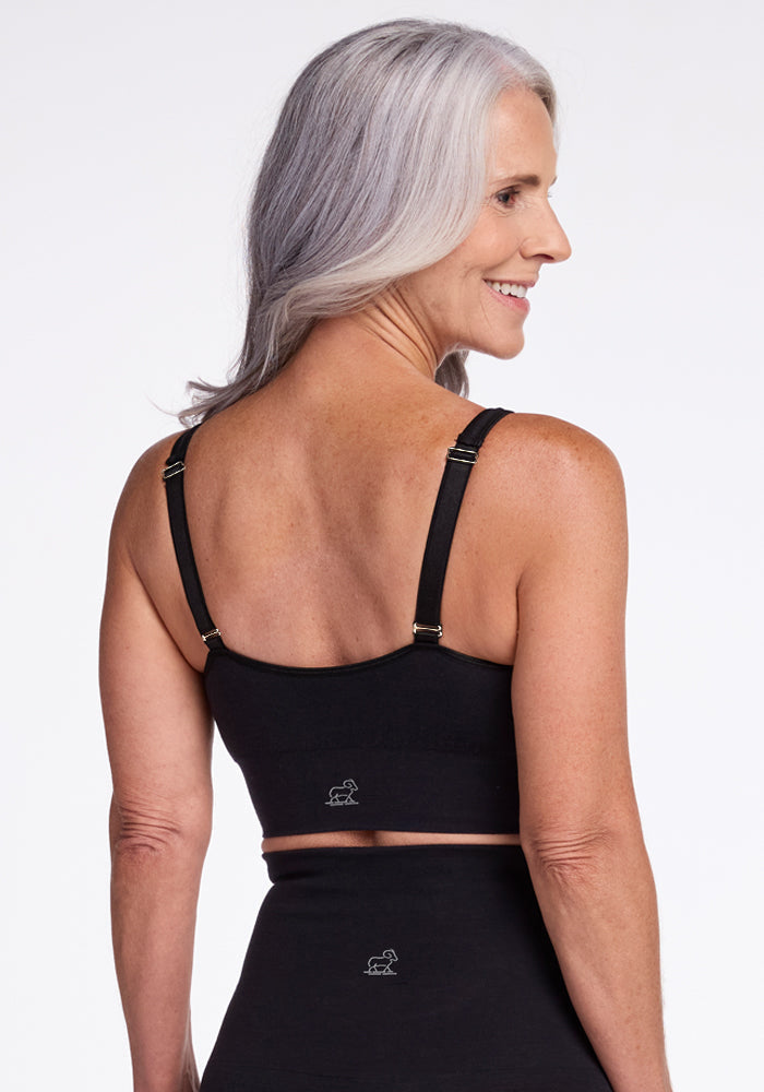 A woman with long gray hair is smiling and facing away from the camera, showcasing the back of a luxurious Woolx Hayden Bra - Black she is wearing. The bra features adjustable straps with metallic clasps. She also appears to be wearing matching black bottoms. The background is plain white.