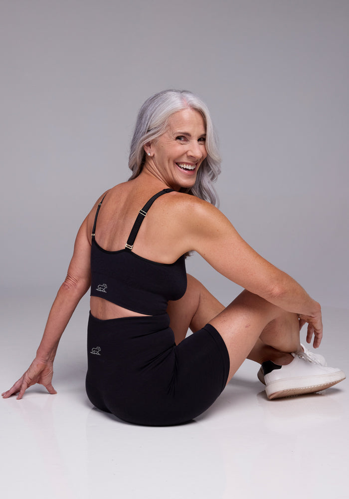 A woman with long white hair is smiling and sitting on the floor, wearing the comfortable Woolx Hayden Bra in black along with matching shorts paired with white sneakers. The image has a plain, light gray background.