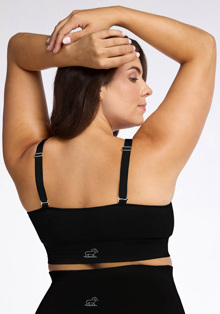 A woman with long brown hair is seen from the back, stretching her arms behind her head. She is wearing a Woolx Hayden Bra - Black with adjustable straps and a small logo near the bottom, designed for comfortable support. The background is plain white.