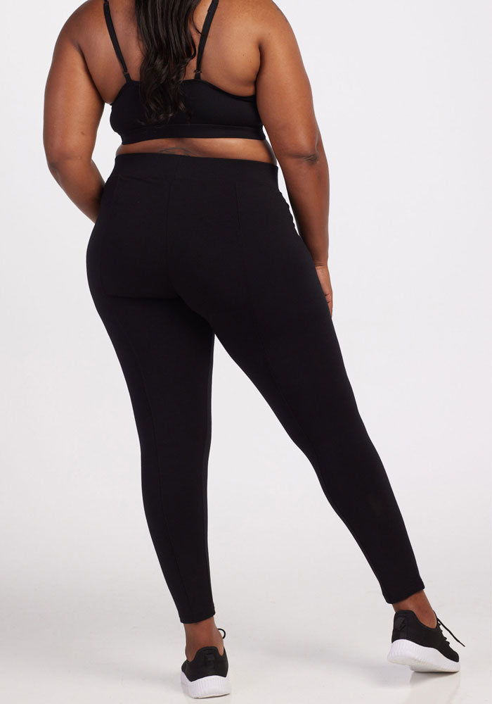 A person is standing against a plain white background, wearing the Woolx Stella Leggings in black that feature flattering back seams, paired with a black sports bra and white and black sneakers.
