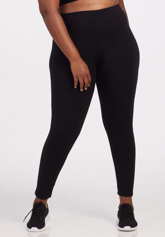 A person wearing the Woolx Stella Leggings - Black, featuring flattering back seams, along with black sneakers stands against a plain background, their left hand resting casually on their thigh. 