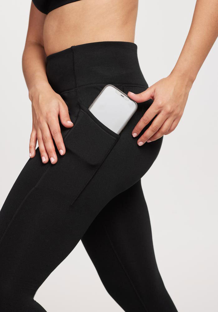 A person wearing Woolx Merino Flex® McKenna Tall leggings with side pockets is placing a smartphone into the pocket.