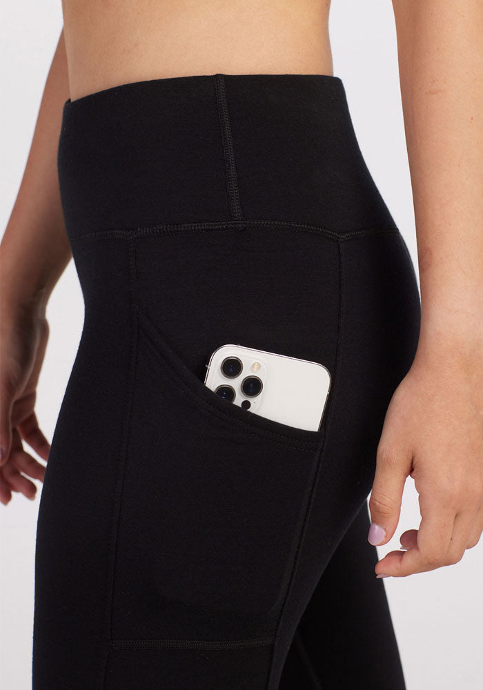 A petite woman stands against a plain background, wearing the Piper Pocket Petite in Black from Woolx. These high-waisted leggings feature a smartphone partially visible in the side pocket and offer cold-weather warmth without compromising on style, thanks to their versatile Merino wool fabric.
