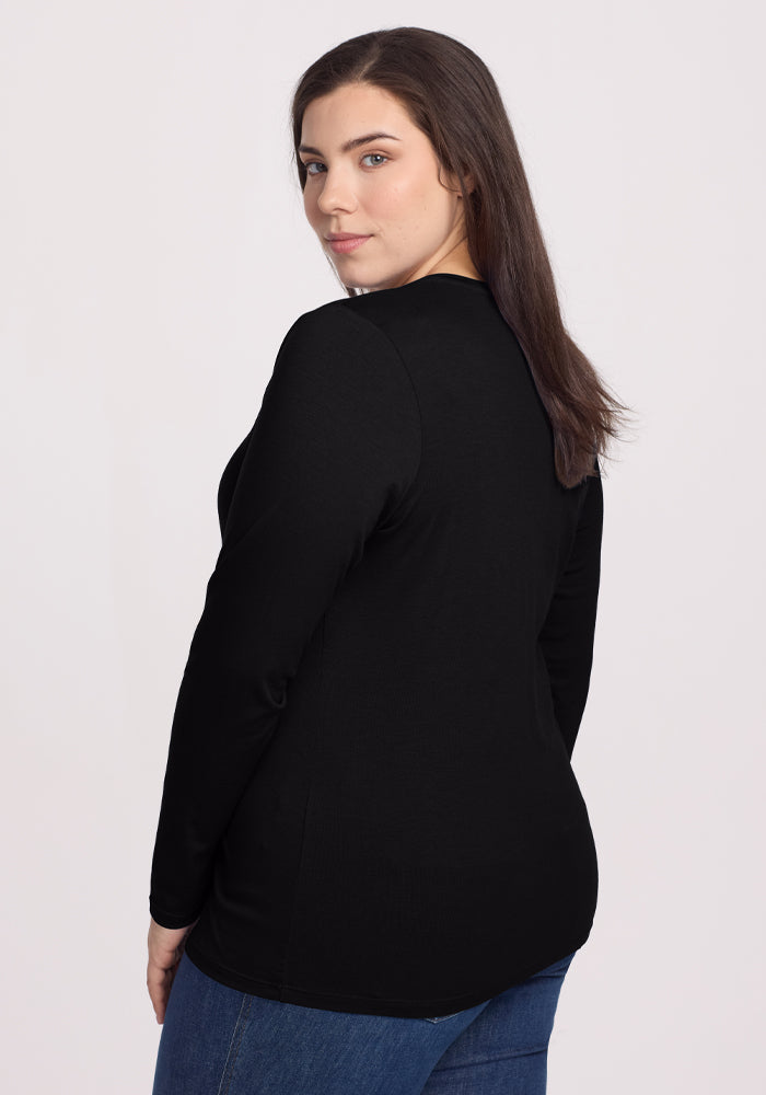 A person with long brown hair stands, glancing over their shoulder, wearing the black Woolx Millie Long Sleeve and blue jeans against a plain white background.