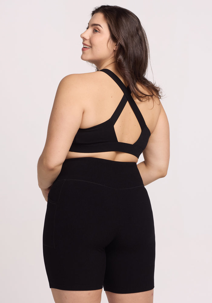 A woman is smiling over her shoulder, wearing the Woolx Miley Bike Shorts in black with a black sports bra. The photo captures the back design of the bra against a plain white background.