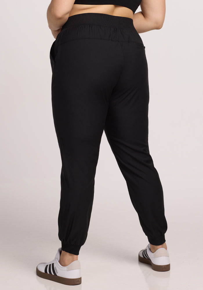 Rear view of a person wearing Woolx's Palmer Joggers in black, made from comfortable Merino fabric, paired with white sneakers featuring black stripes. The plain background highlights their stylish look as they stand slightly angled to the left, showcasing the joggers' deep pockets.