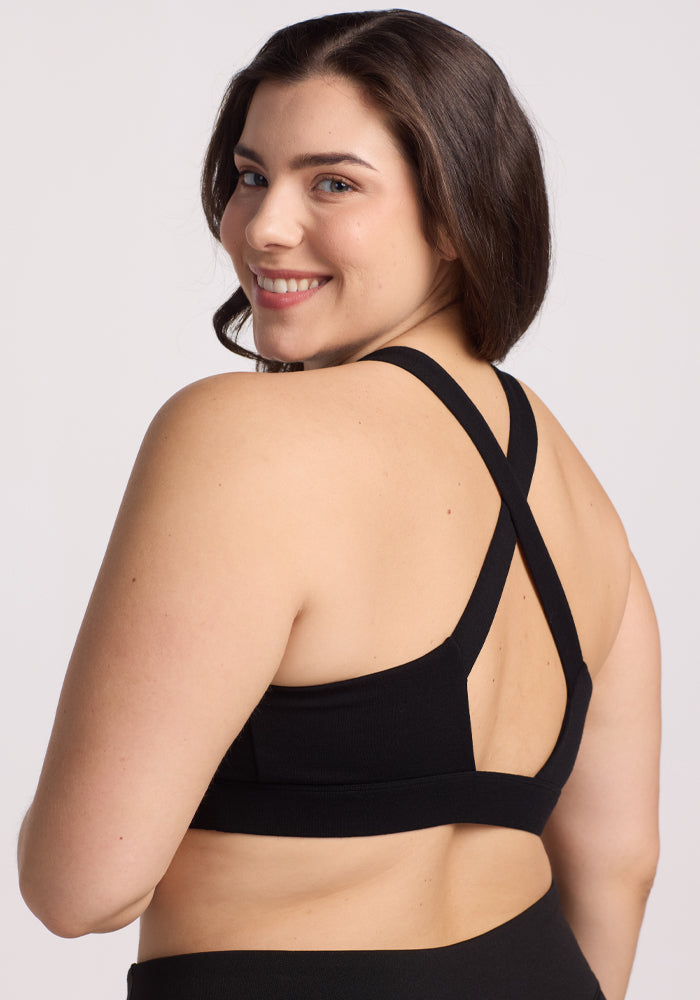 A person with long dark hair smiles over their shoulder, wearing a black Livi Bra by Woolx, featuring a crisscross back and temperature-regulating fabric against a plain, light background.