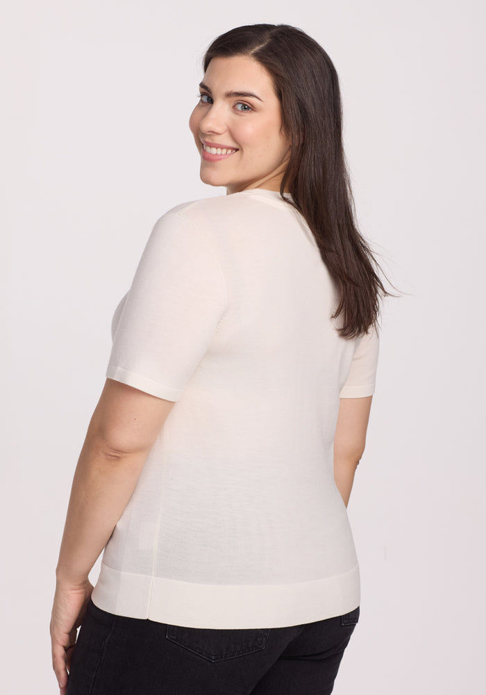 A person with long dark hair smiles over their shoulder, wearing the Woolx Anna Crew Sweater in Dewdrop Pearl, paired with black pants against a plain white background.