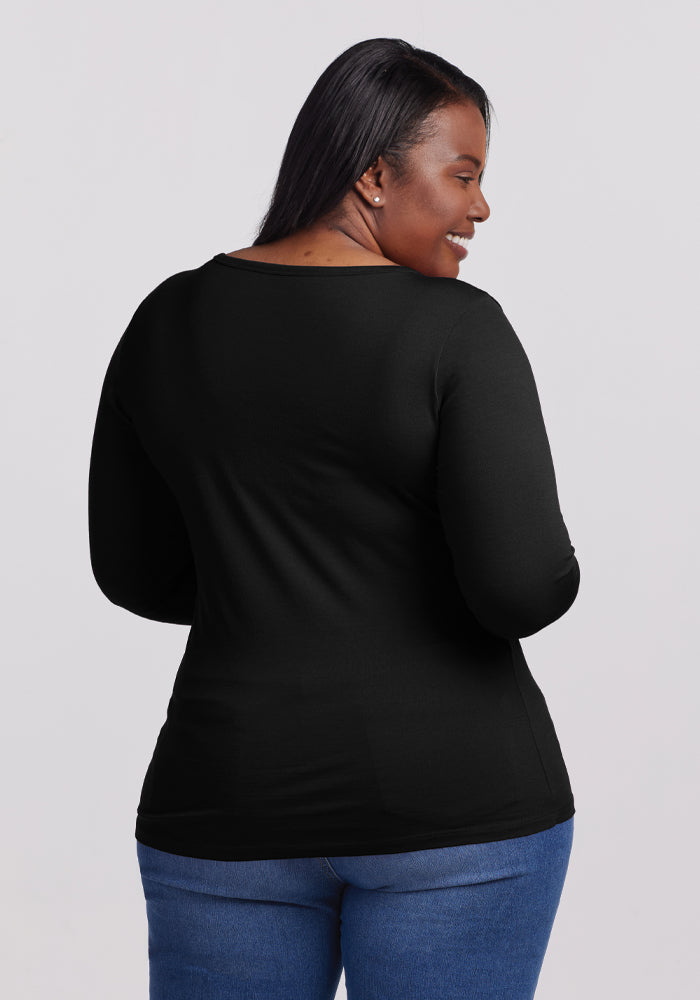 A person with long hair, wearing the Woolx Layla V Neck Top in black and blue jeans, smiles while looking over their shoulder. The simple backdrop adds a touch of elegance to the overall composition.