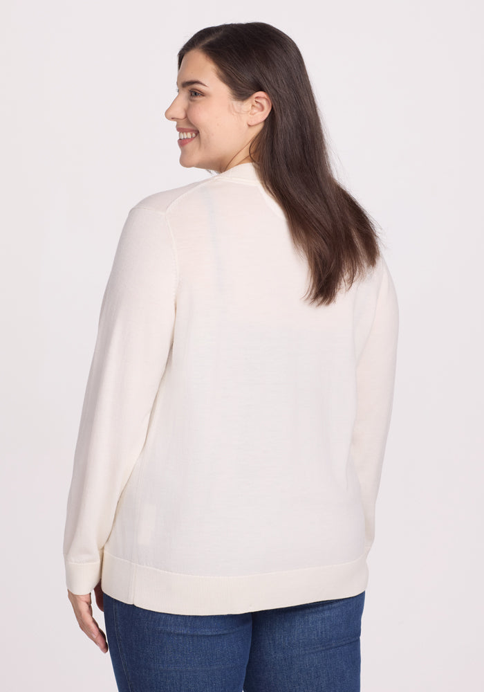 A person with long brown hair, wearing a Woolx Gia Sweater in Dewdrop Pearl and blue jeans, is turned slightly to the side, smiling. The lightweight comfort of the outfit complements the plain white background.