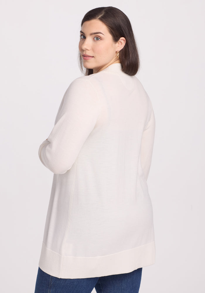 A woman with long dark hair looks over her shoulder at the camera. She's wearing a Woolx Macie Sweater in Dewdrop Pearl and blue jeans, set against a plain white background.