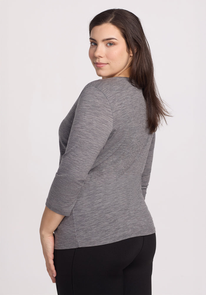 A woman with long brown hair glances over her shoulder at the camera, dressed in a Woolx Elena 3/4 Sleeve V Neck in Graphite Heather and black pants. Made from ultra-fine Merino wool, her outfit stands out against the plain white background, exuding a relaxed fit and effortless style.