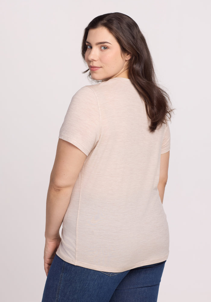 A person with long brown hair stands against a plain white background, turned slightly to the side and looking at the camera. They are wearing a Woolx Liza Crew Neck Tee in Cream Heather and blue jeans.