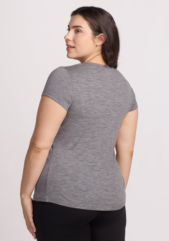 A woman with long brown hair models the Woolx Addie Crew Neck in Graphite Heather, wearing black pants and standing slightly turned to the side, looking over her shoulder against a plain light background.