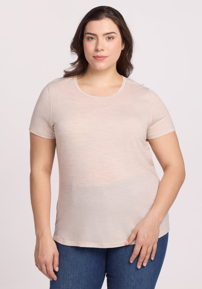 A person with shoulder-length brown hair, wearing a Woolx Liza Crew Neck Tee in Cream Heather and blue jeans, stands against a plain white background, looking at the camera with a neutral expression. 