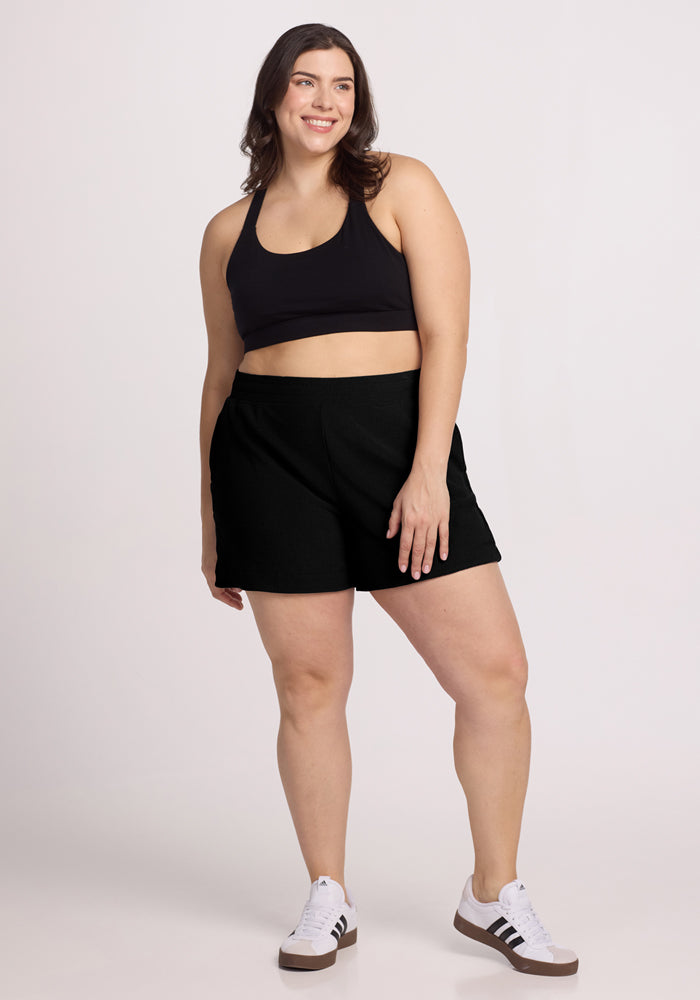 A woman stands smiling, wearing the Maisey Ribbed Shorts in black and a matching sports bra. She has shoulder-length brown hair and white sneakers with black stripes against a plain white background. Her outfit is by Woolx.