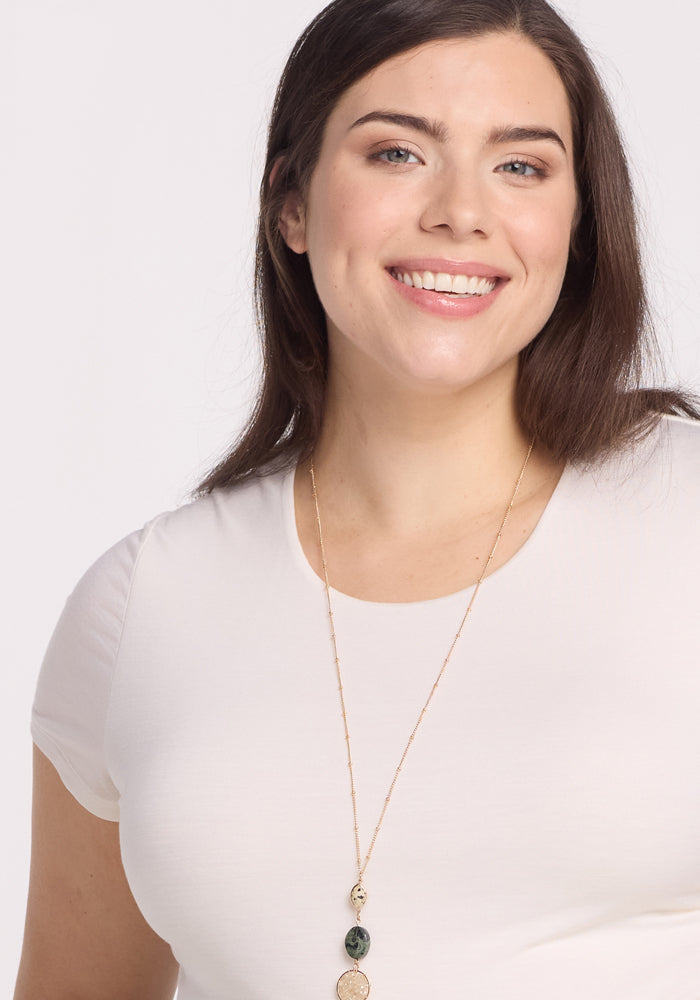 A woman with long brown hair, dressed in the Woolx Gracie Bodysuit - Vanilla, smiles at the camera against a plain light background, accessorized with a gold necklace featuring multiple pendants.