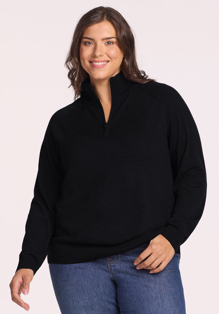 A woman with medium-length brown hair smiles while wearing the Woolx Collins Half Zip Sweater in black and blue jeans, standing against a plain light background. 