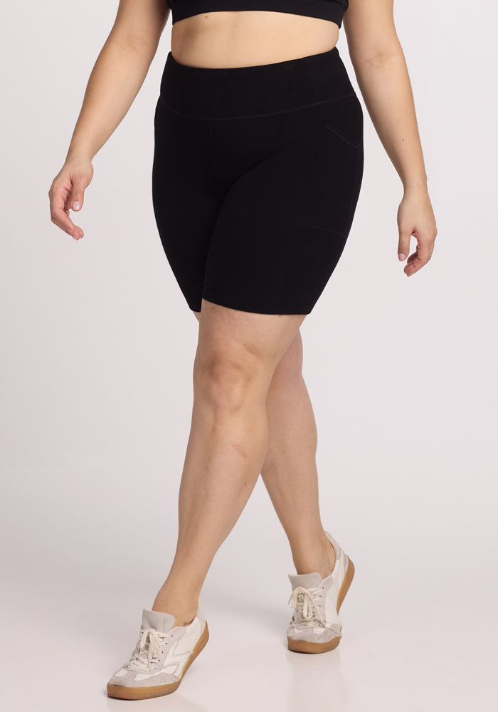 A person in Woolx's Miley Bike Shorts - Black and a matching sports bra stands against a plain background, wearing white sneakers with light tan soles, casually stepping forward. 