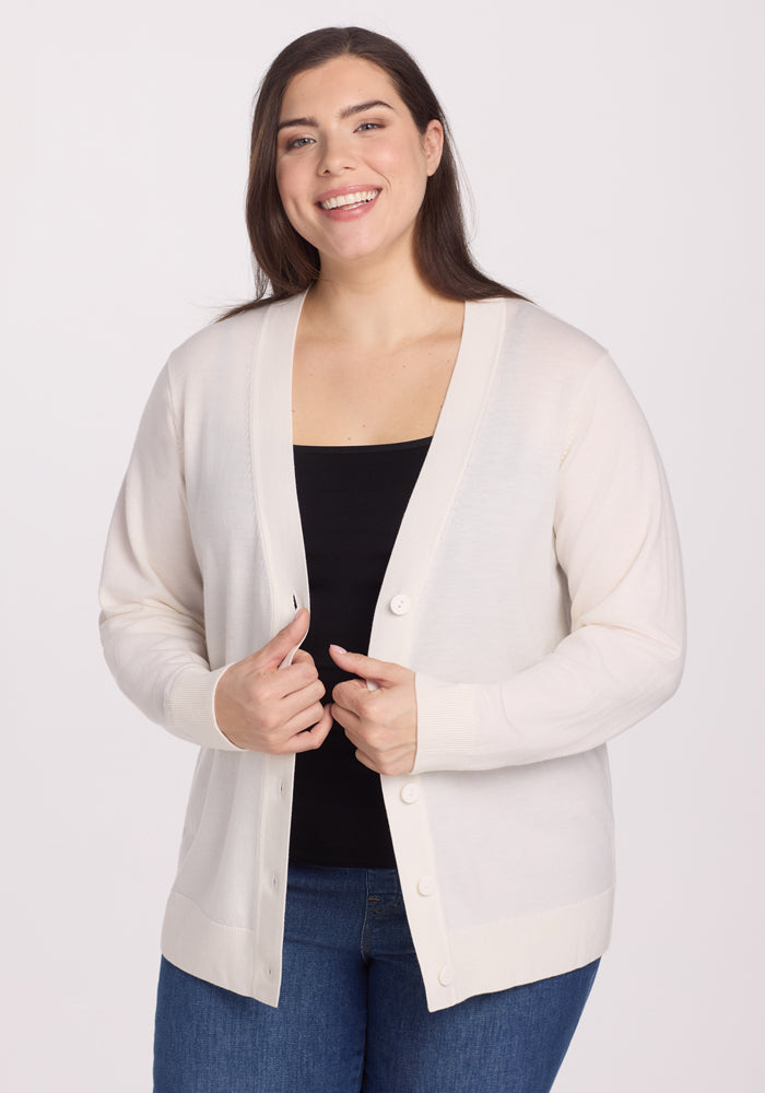 A person smiles while wearing the Woolx Gia Sweater in Dewdrop Pearl, made of odor-resistant Merino wool, over a black top and blue jeans. They stand against a plain white background, holding the lightweight cardigan's edges together. 