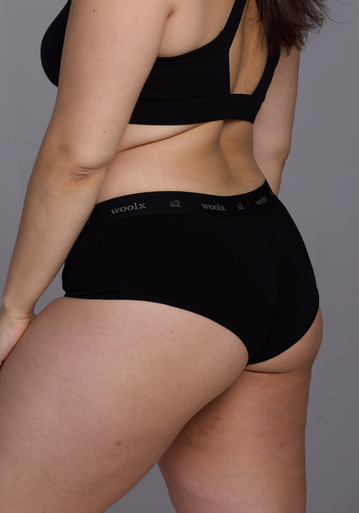 A person poses with their back to the camera, highlighting their side, wearing Woolx's black Lila Boy Shorts. The lightweight fabric offers all-day comfort against a plain gray background.