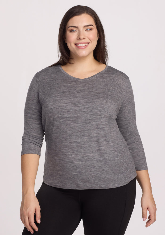 A woman smiles, wearing a Woolx Elena 3/4 Sleeve V Neck in Graphite Heather with black pants, against a light background. The relaxed fit complements her easy stance, arms resting naturally at her sides. 