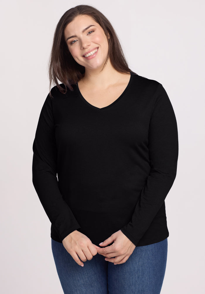 A person with long brown hair smiles wearing the Woolx Millie Long Sleeve - Black and blue jeans, standing against a plain light background, effortlessly combining comfort and style. 