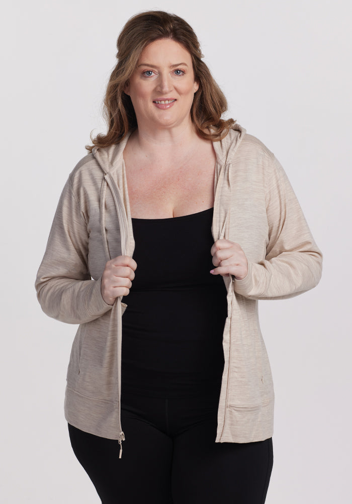 A smiling person with wavy hair stands against a plain background, wearing Woolx's Ryann Hoodie in Cream Heather over a black top and pants. They hold the hoodie open with both hands, highlighting its cozy yet breathable design. 