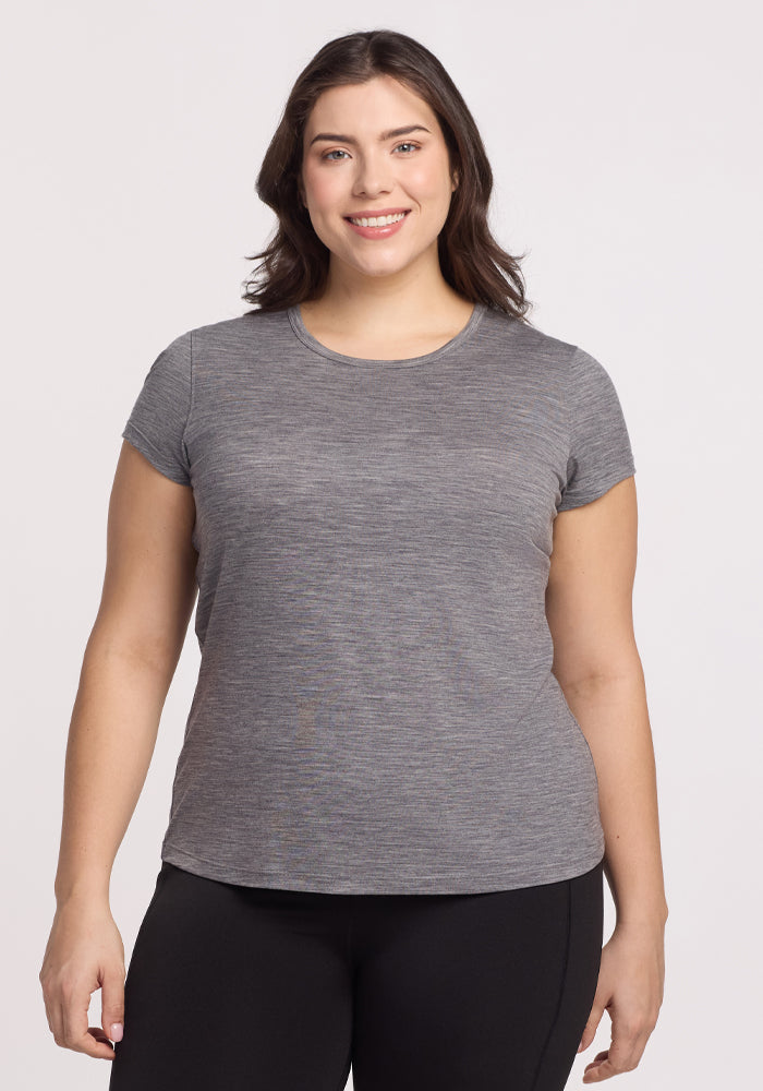 A woman with long dark hair is smiling and wearing the Woolx Addie Crew Neck in Graphite Heather, paired with black pants, standing against a plain light background. 