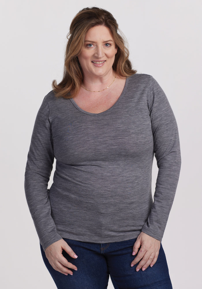 A person with long, light brown hair smiles at the camera, wearing the Woolx Layla V Neck Top in Graphite Heather and blue jeans, standing against a plain white background. 