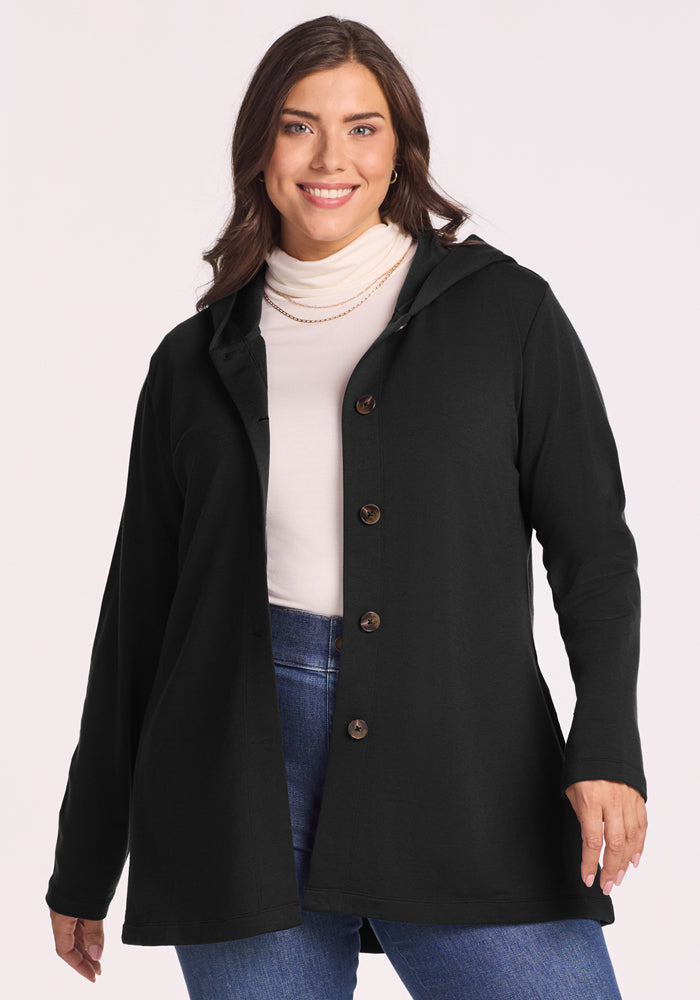A woman with long brown hair, wearing a white turtleneck, blue jeans, and the Woolx Edna Hooded Cardigan in black, smiles while posing against a plain light background. 