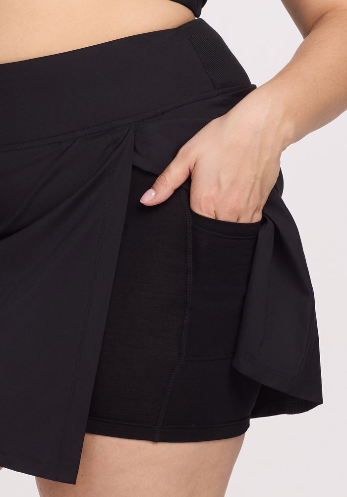 A person is shown wearing a Woolx Topanga Skort in black. The skort features a side slit that reveals shorts underneath, and they are holding their hand in the shorts pocket against a plain white background.