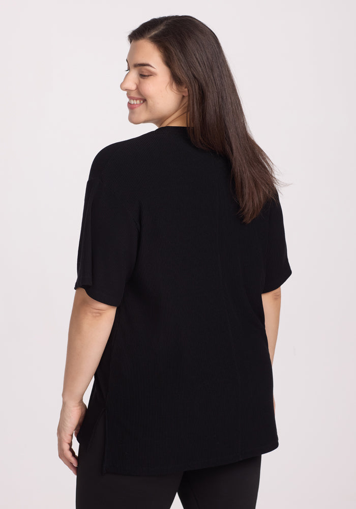 A woman with straight brown hair smiles over her shoulder, wearing the Woolx Vera Ribbed Tunic in black— a relaxed fit, moisture-wicking short-sleeve top—paired with black pants against a plain white background.