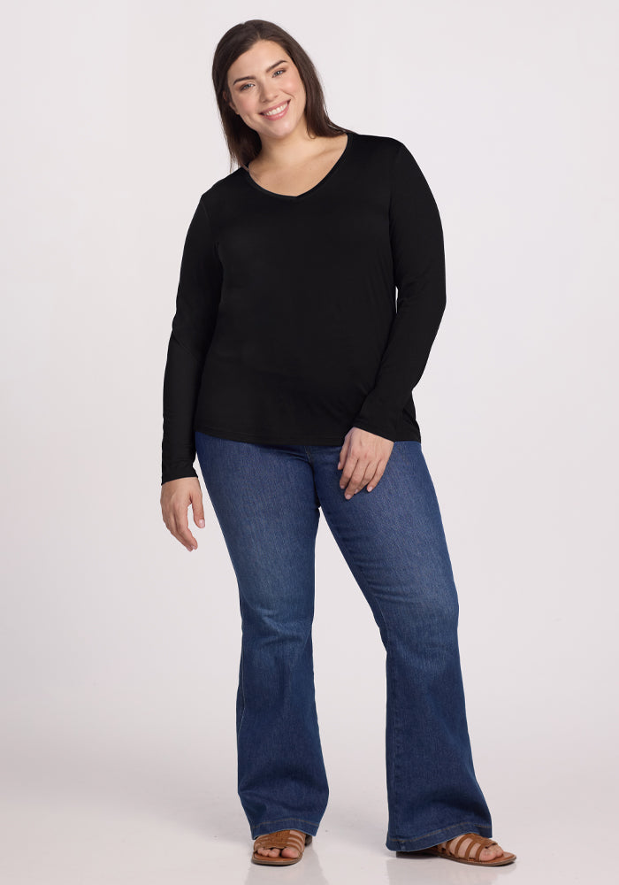 A person with long hair smiles against a light background, wearing a Millie Long Sleeve in black by Woolx, blue jeans, and moisture-wicking sandals.