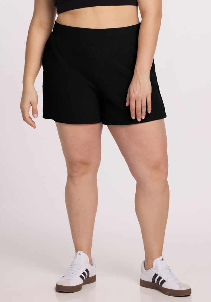 Standing against a plain background, a person models the lower half of their body wearing Woolx's Maisey Ribbed Shorts in black paired with white sneakers featuring black stripes. 