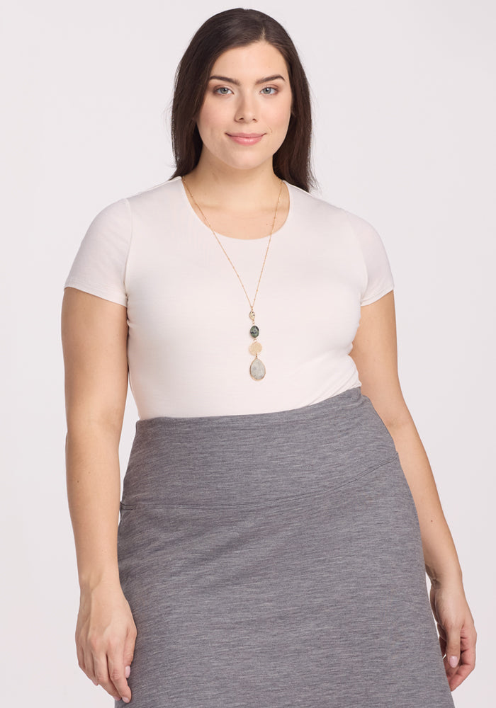 A woman with long brown hair wears the Woolx Gracie Bodysuit in Vanilla and a high-waisted gray skirt, facing the camera against a plain light background to showcase its moisture-wicking, temperature-regulating comfort. 