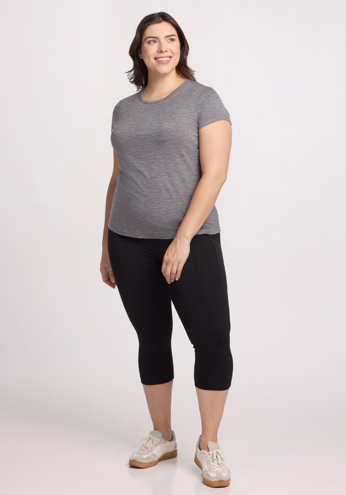 A woman with shoulder-length brown hair is standing and smiling, wearing the Woolx Addie Crew Neck in Graphite Heather, black leggings, and white sneakers against a plain white background.