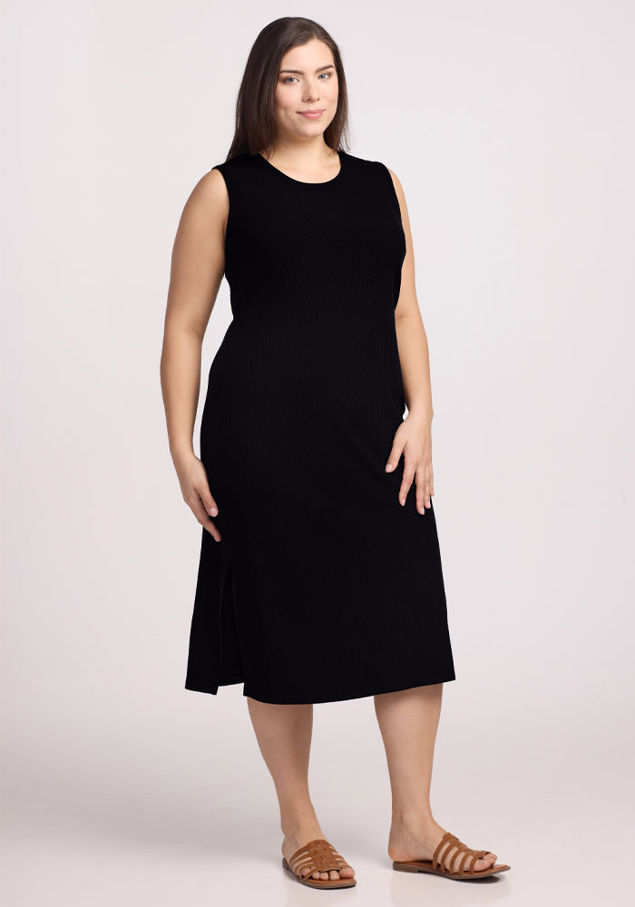 A person stands against a plain background in an Iris Ribbed Dress - Black from Woolx, paired with brown sandals. Their hair is straight and loose, and they have a relaxed expression. 
