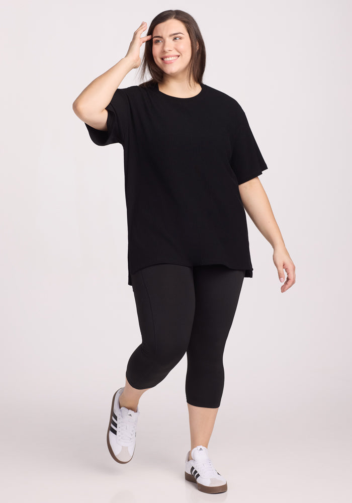 A woman in the Woolx Vera Ribbed Tunic - Black smiles as she walks forward, pairing the relaxed fit top with black leggings and white sneakers against a plain light background.